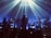 A band is performing on stage with atmospheric lighting. The musicians are playing various instruments including drums and keyboards. Beams of light create a dramatic effect, and the audience is visible as silhouettes in the foreground.