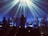 A band is performing on stage with atmospheric lighting. The musicians are playing various instruments including drums and keyboards. Beams of light create a dramatic effect, and the audience is visible as silhouettes in the foreground.