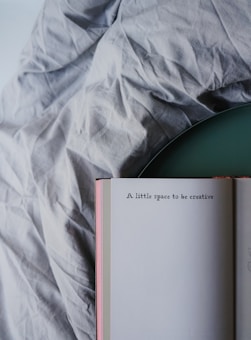 An open book rests on a wrinkled fabric surface with the visible page showing the text 'A little space to be creative'. The book has a light-colored cover and is positioned at an angle.