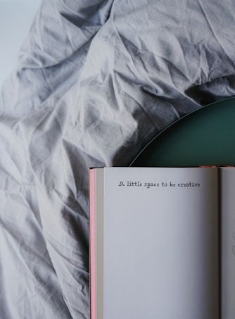 An open book rests on a wrinkled fabric surface with the visible page showing the text 'A little space to be creative'. The book has a light-colored cover and is positioned at an angle.