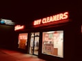 A dry cleaners storefront is brightly lit with neon signs. The red neon lights spell out 'Laundry Cleaners,' while the windows display additional signage promoting services and prices, such as 'Shirts $1.25.' The image is taken at night, providing a stark contrast between the warm glow of the signs and the surrounding darkness.
