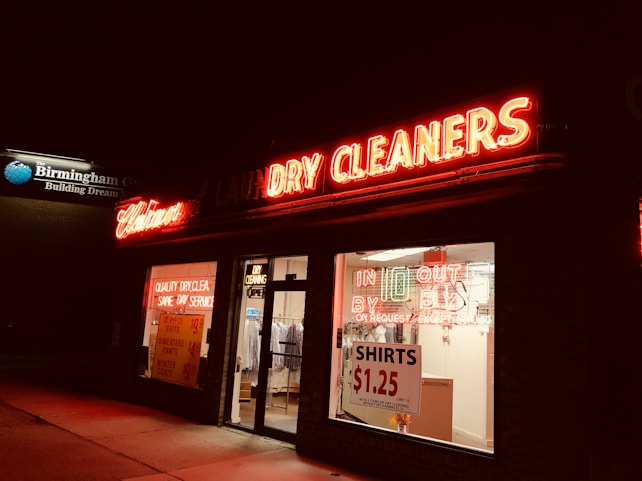 A dry cleaners storefront is brightly lit with neon signs. The red neon lights spell out 'Laundry Cleaners,' while the windows display additional signage promoting services and prices, such as 'Shirts $1.25.' The image is taken at night, providing a stark contrast between the warm glow of the signs and the surrounding darkness.