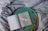 An open book with a simple, artistic illustration of a bird in the middle of one page, placed on a round, green tray. The text on the opposite page reads 'A little space to be creative'. Dried oak leaves are scattered on the tray, which rests on a softly wrinkled, grey fabric.