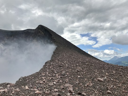 A rugged volcanic landscape with a steep, rocky slope leading to the crater. Thick clouds of smoke or steam are billowing out from the crater, merging with a layer of scattered clouds in the sky. The horizon reveals distant mountains under a partly cloudy sky.
