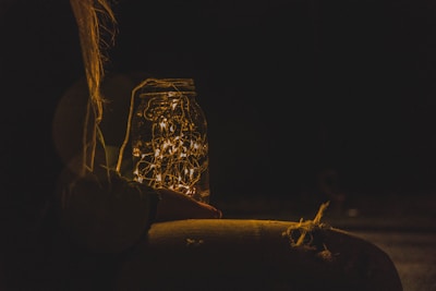 A hand holds a jar filled with glowing fairy lights in a dark setting. The scene is dimly illuminated by the warm, yellow light emitted from the jar, creating a cozy and magical atmosphere. The soft glow emphasizes the contrast between light and shadow.