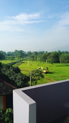 A lush, green landscape features a field of grass with scattered trees in the background under a partly cloudy sky. In the mid-ground, there's a yellow excavator parked, suggesting ongoing development or construction activities. The foreground includes a modern, white building corner with a sloped roof of another structure visible.