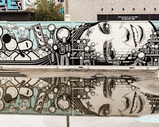 A vibrant mural features a close-up of a smiling woman's face, surrounded by stylized cartoon-like characters with geometric shapes and patterns. The mural reflects in a puddle on the ground, creating a symmetrical view. The background includes hints of greenery and other graffiti artworks.