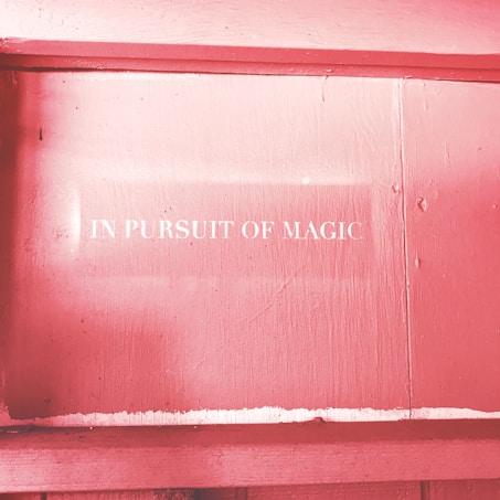 White text reading 'IN PURSUIT OF MAGIC' is displayed on a textured pink surface, possibly a painted wooden wall. The lighting creates a soft, warm atmosphere, enhancing the dreamy feel of the message.