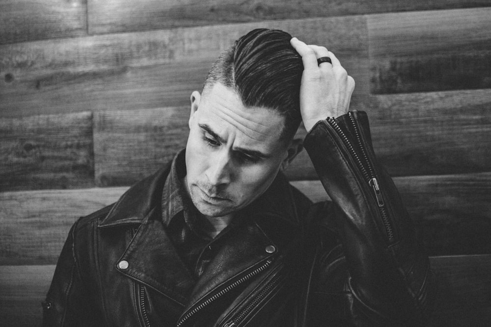 grayscale photography of man wearing leather jacket leaning against wall while fixing hair