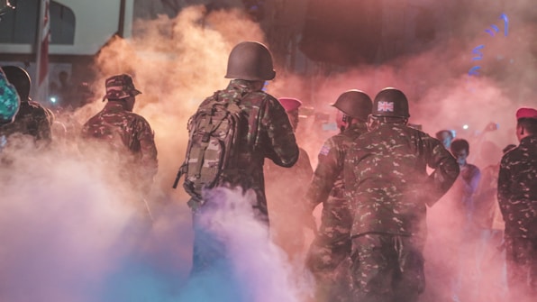 A group of soldiers wearing camouflage uniforms and helmets are enveloped in colorful smoke. The scene is likely during a military exercise or a controlled environment. The atmosphere is intense and active, with varying hues of orange, pink, and blue smoke swirling around.