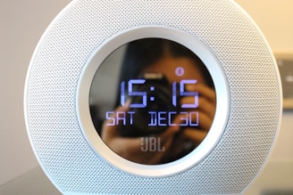 A digital clock with a circular design displays the time as 15:15 and the date as Saturday, December 30. The clock has a perforated metallic outer casing with a reflective surface, visible due to the blurred reflection of a person with a camera taking a photo.