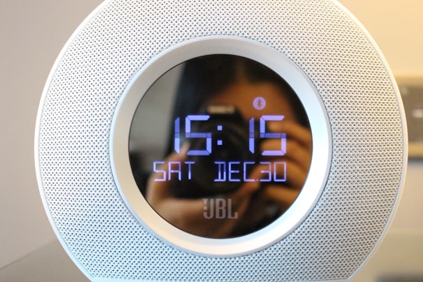 A digital clock with a circular design displays the time as 15:15 and the date as Saturday, December 30. The clock has a perforated metallic outer casing with a reflective surface, visible due to the blurred reflection of a person with a camera taking a photo.