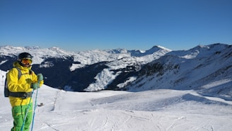 A skier wearing a bright insulated jacket standing against a snowy mountain backdrop at sunrise.