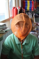 A person is seated indoors wearing a light green shirt and a tan baseball cap. They have an orange net over their face, which is typically used for catching insects or small animals. In the background, there are colorful items like condiment bottles and cleaning brushes on shelves.