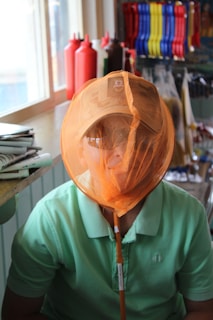 A person is seated indoors wearing a light green shirt and a tan baseball cap. They have an orange net over their face, which is typically used for catching insects or small animals. In the background, there are colorful items like condiment bottles and cleaning brushes on shelves.