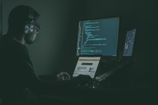 A person sits in a dark room wearing headphones, working on multiple computer screens displaying code. One screen prominently shows programming code, while another laptop screen displays a webpage.
