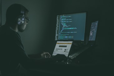 A person sits in a dark room wearing headphones, working on multiple computer screens displaying code. One screen prominently shows programming code, while another laptop screen displays a webpage.