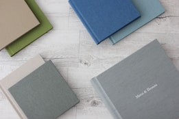 A collection of hardcover books with fabric covers is arranged on a white wooden surface. The books come in various colors, including green, beige, blue, and gray. One book in the bottom right corner features the names 'Marco & Eleonora' embossed in white on the cover.