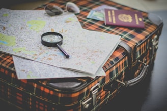 A plaid suitcase is topped with a detailed city map, a magnifying glass for close inspection, a pair of sunglasses, and a passport.