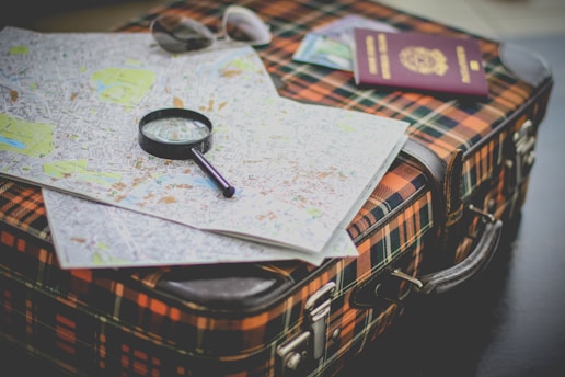 A plaid suitcase is topped with a detailed city map, a magnifying glass for close inspection, a pair of sunglasses, and a passport.
