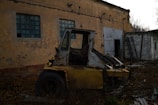 An abandoned and rusted forklift sits amidst overgrown weeds and debris in front of a dilapidated building with peeling paint and dirty, grid-like windows. The atmosphere is desolate, with scattered metal scraps and a leaning ladder against the wall. The scene appears neglected, with dark undertones and a feeling of decay.