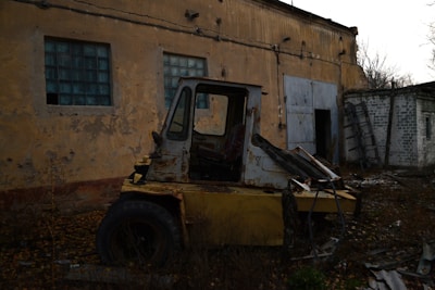 An abandoned and rusted forklift sits amidst overgrown weeds and debris in front of a dilapidated building with peeling paint and dirty, grid-like windows. The atmosphere is desolate, with scattered metal scraps and a leaning ladder against the wall. The scene appears neglected, with dark undertones and a feeling of decay.