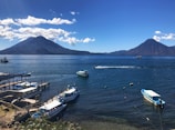 A serene landscape featuring a large lake with a backdrop of two prominent volcanoes under a clear blue sky. Boats are docked along the wooden piers in the foreground, while a few are sailing across the water. The setting exudes a tranquil and picturesque ambiance.