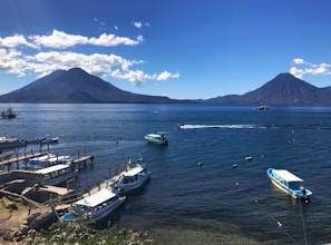 A serene landscape featuring a large lake with a backdrop of two prominent volcanoes under a clear blue sky. Boats are docked along the wooden piers in the foreground, while a few are sailing across the water. The setting exudes a tranquil and picturesque ambiance.