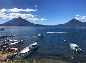 A serene landscape featuring a large lake with a backdrop of two prominent volcanoes under a clear blue sky. Boats are docked along the wooden piers in the foreground, while a few are sailing across the water. The setting exudes a tranquil and picturesque ambiance.