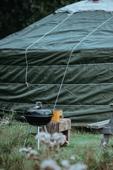 A large green tent is prominently featured with a rounded top and visible ropes securing it. In front of the tent, there is a black charcoal grill next to a wooden bench and a small table holding a yellow radio. The scene is set in a grassy area with some flowers in the foreground.