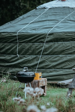 A large green tent is prominently featured with a rounded top and visible ropes securing it. In front of the tent, there is a black charcoal grill next to a wooden bench and a small table holding a yellow radio. The scene is set in a grassy area with some flowers in the foreground.