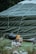 A large green tent is prominently featured with a rounded top and visible ropes securing it. In front of the tent, there is a black charcoal grill next to a wooden bench and a small table holding a yellow radio. The scene is set in a grassy area with some flowers in the foreground.