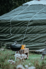 A large green tent is prominently featured with a rounded top and visible ropes securing it. In front of the tent, there is a black charcoal grill next to a wooden bench and a small table holding a yellow radio. The scene is set in a grassy area with some flowers in the foreground.