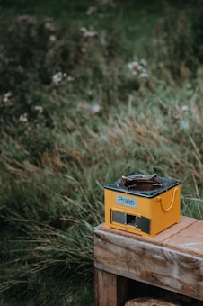 A yellow stove labeled 'Prakti' is placed on a wooden surface. The background consists of tall grass with blurred flowers, suggesting an outdoor setting.