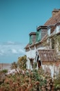 A charming cottage with green window shutters and a tiled roof is surrounded by lush greenery and flowering plants. The sky is clear and blue, adding to the serene rural atmosphere.