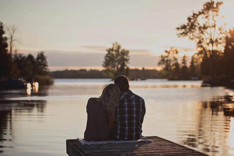 In the image, a couple is sitting on a wooden dock that extends into a serene lake. The man and woman are facing away from the camera, their backs to it. They appear to be enjoying each other's company while taking in the beautiful view of the sunset over the water. The sky above them is painted with hues of orange and yellow, creating a warm and romantic atmosphere. In the background, there are trees lining the lake, adding depth to the scene.