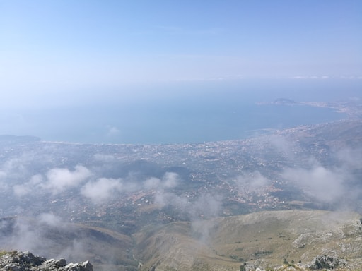 A breathtaking panoramic view from a high vantage point showcasing a vast landscape. The foreground features rocky terrain with patches of greenery, transitioning to rolling hills. The middle ground includes a sprawling urban area by a large bay. Wispy clouds are scattered at different heights, and the distant horizon outlines the calm sea under a mostly clear sky.