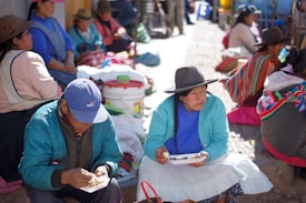 A group of people, predominantly women, are sitting outdoors, eating and conversing. They are wearing traditional clothing, including hats and colorful garments. One woman and man are seated in the foreground eating from plates, while others are grouped along the sides of a narrow, sunlit path. Various containers and items are scattered around them.