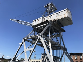 A large, industrial crane structure is positioned against a clear blue sky. The crane has a rusted metal surface and is supported by a triangular framework of metal beams.
