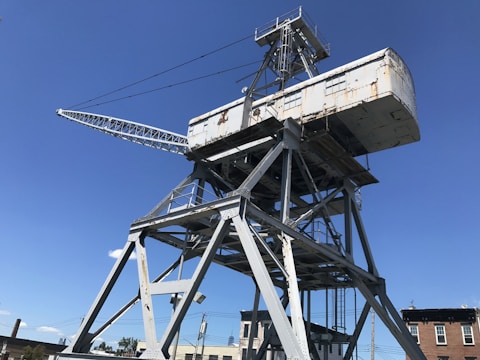 A large, industrial crane structure is positioned against a clear blue sky. The crane has a rusted metal surface and is supported by a triangular framework of metal beams.