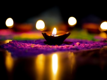 A series of small oil lamps with flames are placed on a colorful surface, likely decorated with rangoli patterns. The background is dark, enhancing the warm glow of the lamps.