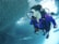 A scuba diver is underwater next to a large school of fish, wearing a purple wetsuit and dive equipment including an air tank and mask. Sunlight filters through the water, creating a serene and vibrant underwater scene.