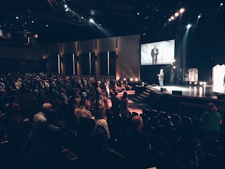A large auditorium filled with many people standing and facing a stage. On the stage, a person is speaking, and a large screen above displays the speaker. The lighting is dim with focused spotlights on the stage area.
