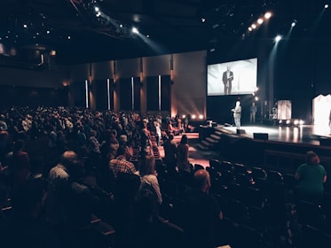 A large auditorium filled with many people standing and facing a stage. On the stage, a person is speaking, and a large screen above displays the speaker. The lighting is dim with focused spotlights on the stage area.
