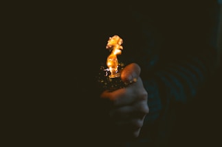 A close-up of a hand holding a lit lighter against a dark background. The flame is small yet bright, casting a warm glow and creating a stark contrast with the surrounding darkness.