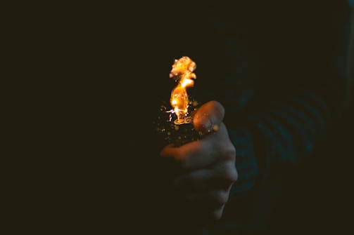 A close-up of a hand holding a lit lighter against a dark background. The flame is small yet bright, casting a warm glow and creating a stark contrast with the surrounding darkness.