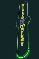 A vertical neon sign with the words 'bistro market' displayed in bright yellow against a dark background. The outline of a shopping cart is integrated into the design above the text, and the sign is surrounded by a glowing green border.