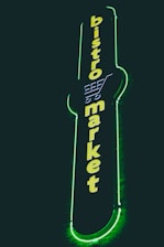 A vertical neon sign with the words 'bistro market' displayed in bright yellow against a dark background. The outline of a shopping cart is integrated into the design above the text, and the sign is surrounded by a glowing green border.