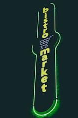 A vertical neon sign with the words 'bistro market' displayed in bright yellow against a dark background. The outline of a shopping cart is integrated into the design above the text, and the sign is surrounded by a glowing green border.