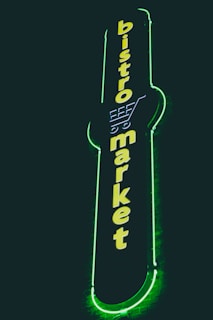 A vertical neon sign with the words 'bistro market' displayed in bright yellow against a dark background. The outline of a shopping cart is integrated into the design above the text, and the sign is surrounded by a glowing green border.